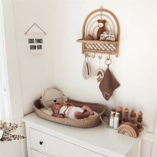 Dans le coin d'une chambre d'enfant aux murs blancs, on voit une commode blanche surmontée d'une corbeille à langer dans laquelle se trouve un nourrisson. Près de lui, tous les indispensables pour le change r, et au-dessus, sur le mur, une étagère en rotin en forme d'arc en ciel est suspendue. L'image fait très instagram.