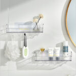 Sur un mur blanc, deux étagères murales rectangulaires transparentes l'une sous l'autre en décalé avec des flacons et des crèmes dedans. Il y a une fleur de douche et une petite râpe accrochées aux crochets du dessous. On aperçoit à droite une partie d'un miroir rond au contour doré.