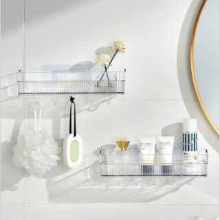 Sur un mur blanc, deux étagères murales rectangulaires transparentes l'une sous l'autre en décalé avec des flacons et des crèmes dedans. Il y a une fleur de douche et une petite râpe accrochées aux crochets du dessous. On aperçoit à droite une partie d'un miroir rond au contour doré.
