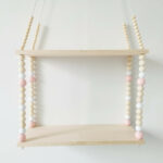 Une étagère en bois naturel suspendue avec 2 tablettes et entre chaque tablette une rangée de perles en bois roses et blanches.