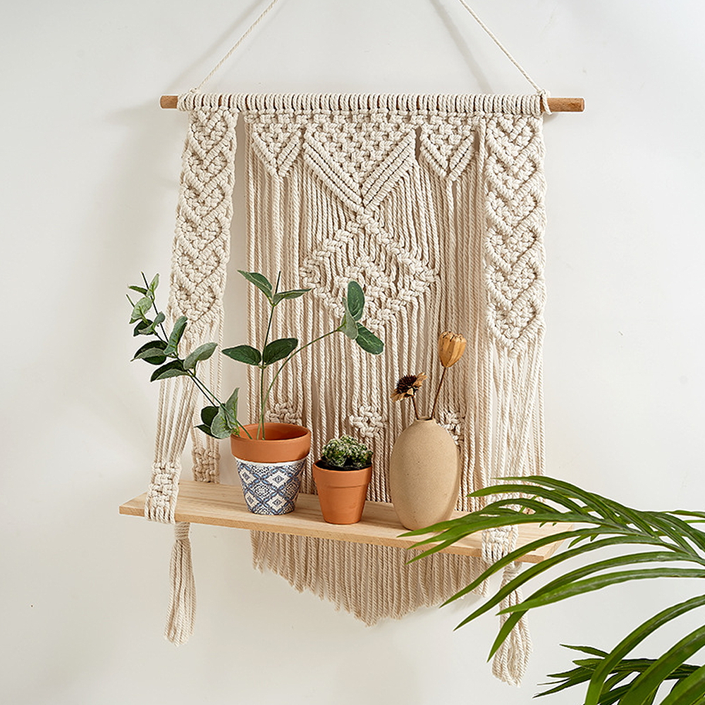 Étagère murale avec des cordes en coton beige et un support en bois où sont posés des pots de fleurs. Une feuille de palmier à droite. Sur mur blanc.