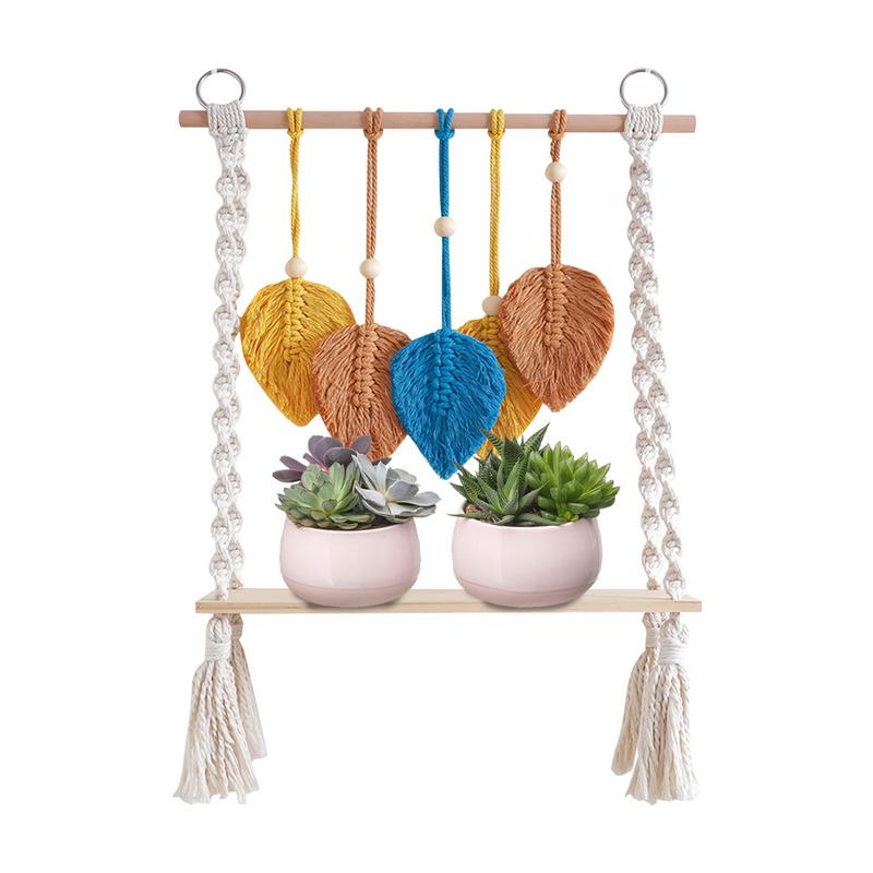 étagère murale avec cordes beige de chaque côté et support en bois comme une balançoire. Au-dessus, des feuilles orange et bleus en macramé. sur le support en bois 2 plantes. Sur fond blanc.