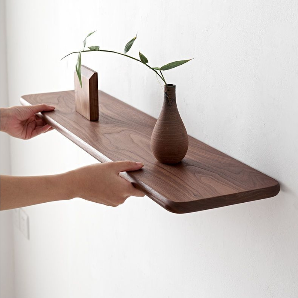 Étagère murale en bois foncé et vernis, de forme rectangulaire fixée sur un mur blanc. Dessus un vase marron avec une branche verte et un objet carré du même marron. Deux mains tiennent le devant de l'étagère.