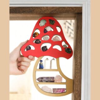 Étagère murale en forme de champignon en bois avec le pied beige et le chapeau rouge. L'ensemble est pourvu de trous dans lesquels sont stockés des cristaux.Une main tient l'étagère par le coin gauche du chapeau du champignon. L'étagère se trouve dans un angle d'encadrement de porte en bois.