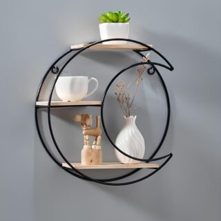 Étagère murale en forme de demi-lune en métal noir. Elle dispose de 3 petites étagères en bois. Tout en haut, une petite plante verte dans un pot blanc, le niveau du milieu une tasse blanche, en bas, un vase blanc avec une fleur sèche, un sujet en bois. Fixée sur un mur gris.