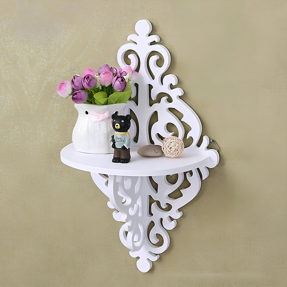 Étagère murale blanche en forme de losange avec des arabesques. Une tablette blanche sur laquelle sont posés un pot blanc avec des fleurs et des objets de décoration. Fixée sur un mur beige.