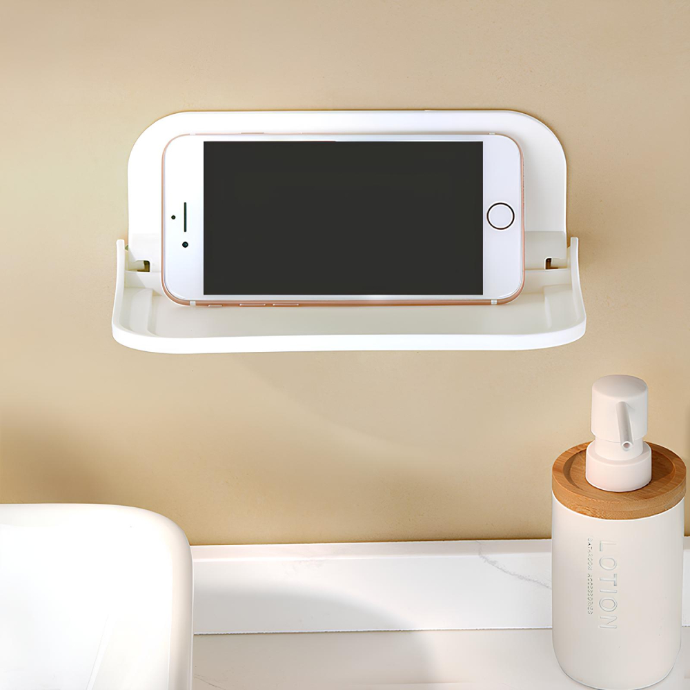 Étagère murale en plastique blanc pliante. Elle est ouverte et un téléphone y est posé en orientation paysage. Elle est fixée sur un mur beige. En bas à droite, un flacon blanc et marron, à gauche, le coin du lavabo.