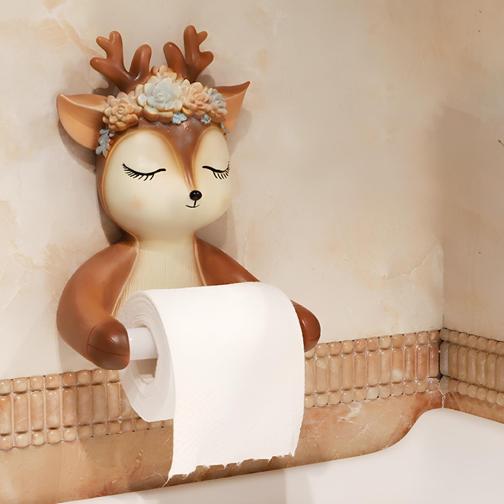 Porte-papier toilette en forme de cerf enfantin avec couronne de fleurs et les yeux fermés qui tient entre ses pattes un rouleau blanc. Fixée sur un mur beige avec une bande plus foncée en dessous.