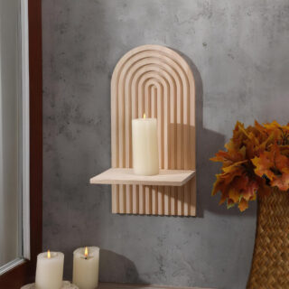 Étagère murale en bois dans le style bohème en forme d'arc-en-ciel avec un support rectangulaire. Une bougie beige allumée est posée dessus. L'étagère est fixée sur un mur gris. Deux bougies blanches allumées à gauche, des feuilles d'automne à droite dans un vase en rotin.